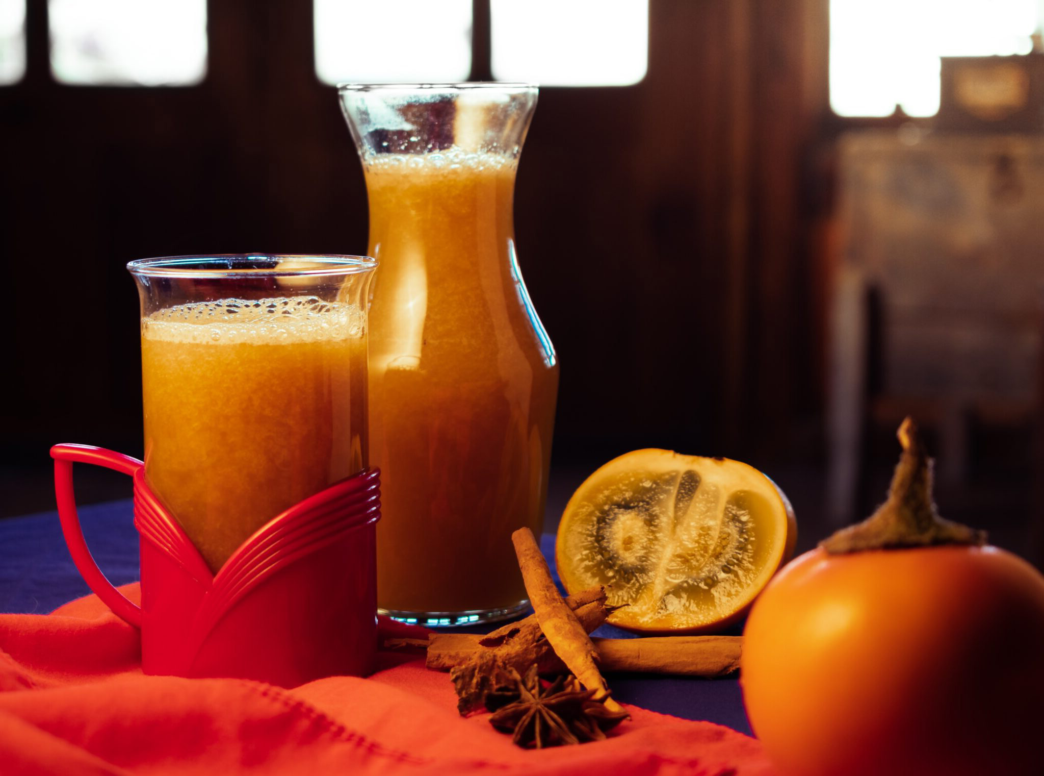 Tazza di vetro con manico rosso e caraffa di Canelazo, una bevanda tradizionale ecuadoregna a base di aguardiente, cannella e agrumi, servita calda. Accanto, spezie come cannella e anice stellato, oltre a un agrume tagliato e un cachi. L'atmosfera è calda e accogliente, con un'illuminazione soffusa che richiama un ambiente rustico