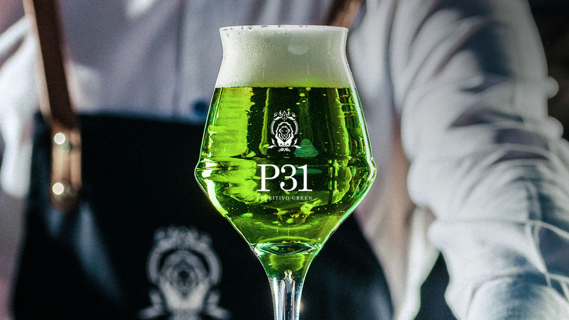 Calice elegante con P31 Green Beer (P31 Aperitivo Green più una birra bionda), una bevanda dal colore verde intenso e brillante con una schiuma leggera. Sullo sfondo, un bartender in camicia bianca e grembiule scuro con dettagli dorati, che conferisce un'atmosfera sofisticata e raffinata